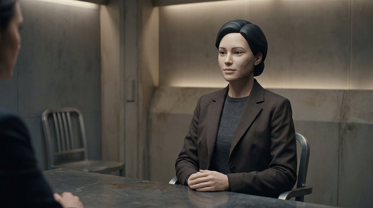 Lucene-9000, a humanoid machine of female form in a dark jacket, seated to one side at a bare interview-room table with her hands folded, watching someone whose shoulder shows in the foreground. Her matte face carries a fine seam at the jaw and temple, her eyes are recessed lenses, and her collar carries no rank mark.