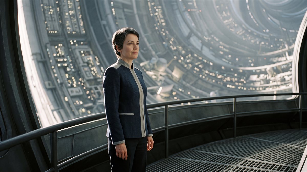 Space Commissioner Zara Wayland on an upper walkway inside Eden Space Habitat, a woman in a dark suit standing squarely with her hands at her sides, facing the viewer. The habitat's inhabited interior curves away and upward behind her, lit along its length.