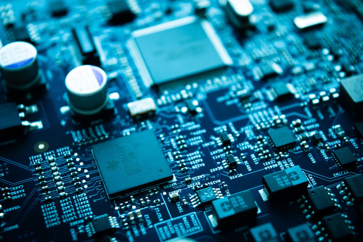 A close-up of a densely populated circuit board lit in deep blue: two large square processor chips, cylindrical capacitors standing proud of the surface, and hundreds of small surface-mount components among printed reference codes, the far side of the board falling out of focus.