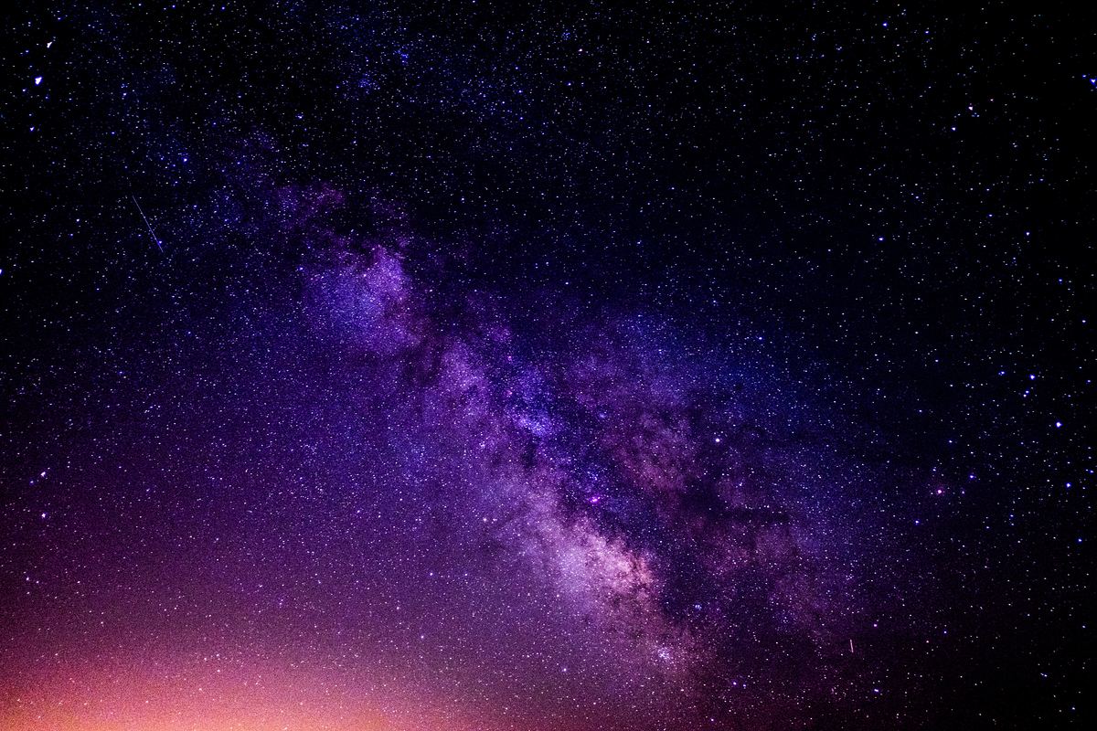 A night-sky photograph of the Milky Way, its band of violet star clouds and dark dust lanes running diagonally across the frame, thousands of individual stars resolved around it, a thin meteor streak at the upper left and a warm orange glow along the horizon at the bottom.