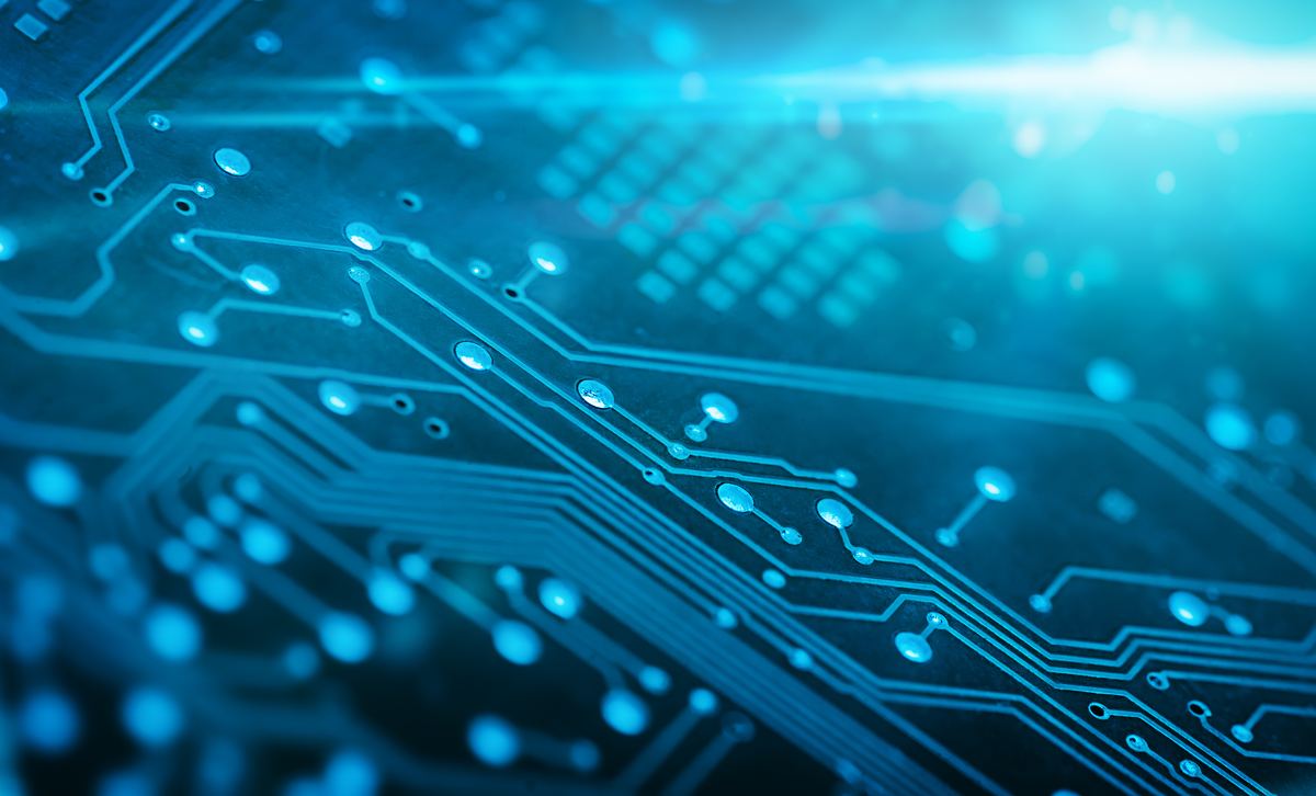 A macro view along a circuit board, its etched traces and solder points glowing cyan against a dark surface and running diagonally out of frame toward a bright flare of light in the corner. No components, only the pathways.
