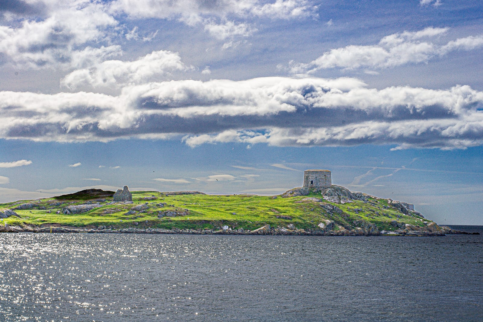 A low green island across dark open water under a heavy shelf of cloud: bare rock at the tideline, grass above it, a round stone watchtower on the summit and the gabled ruin of a small stone house standing further along the ridge.