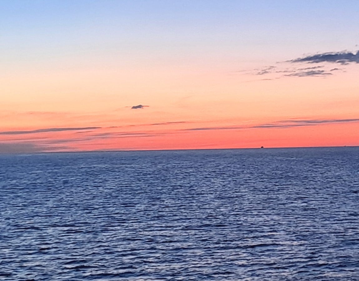 Open ocean at dusk with no land anywhere in sight: violet water running out to a band of pink light on the horizon, and two distant vessels hull-down on the line between them.
