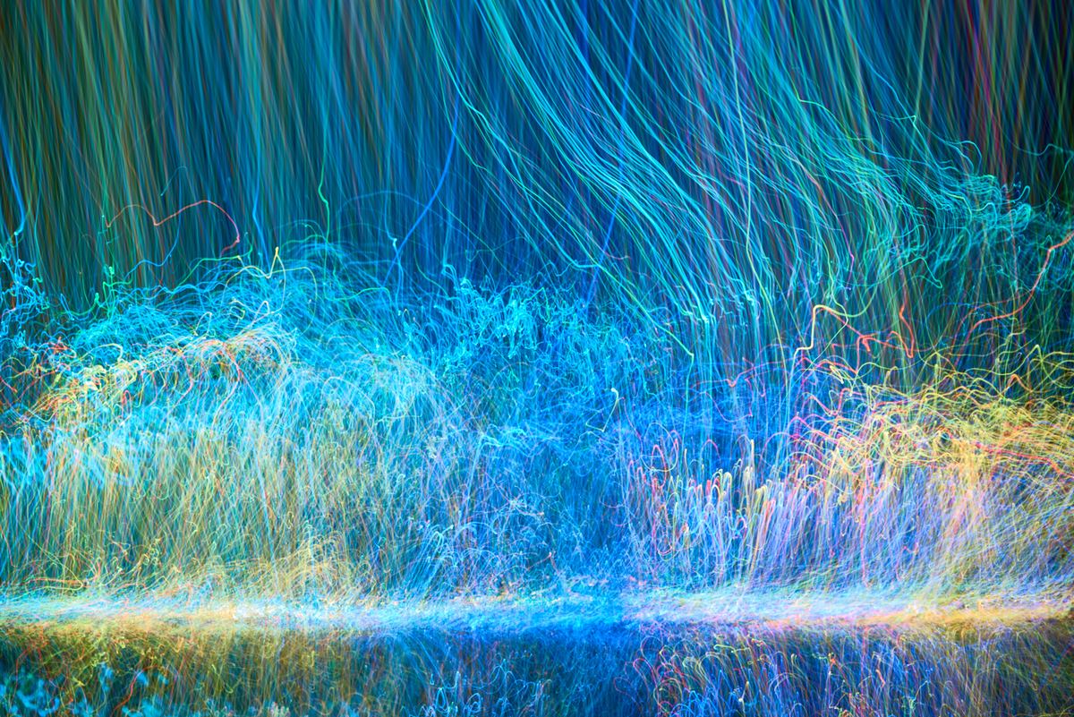 A dense tangle of multicoloured light trails, as though a long exposure had been swept through a field of lights: cyan and blue threads dominating, shot through with green, yellow and orange, dark at the top and massing into a bright band across the lower half.
