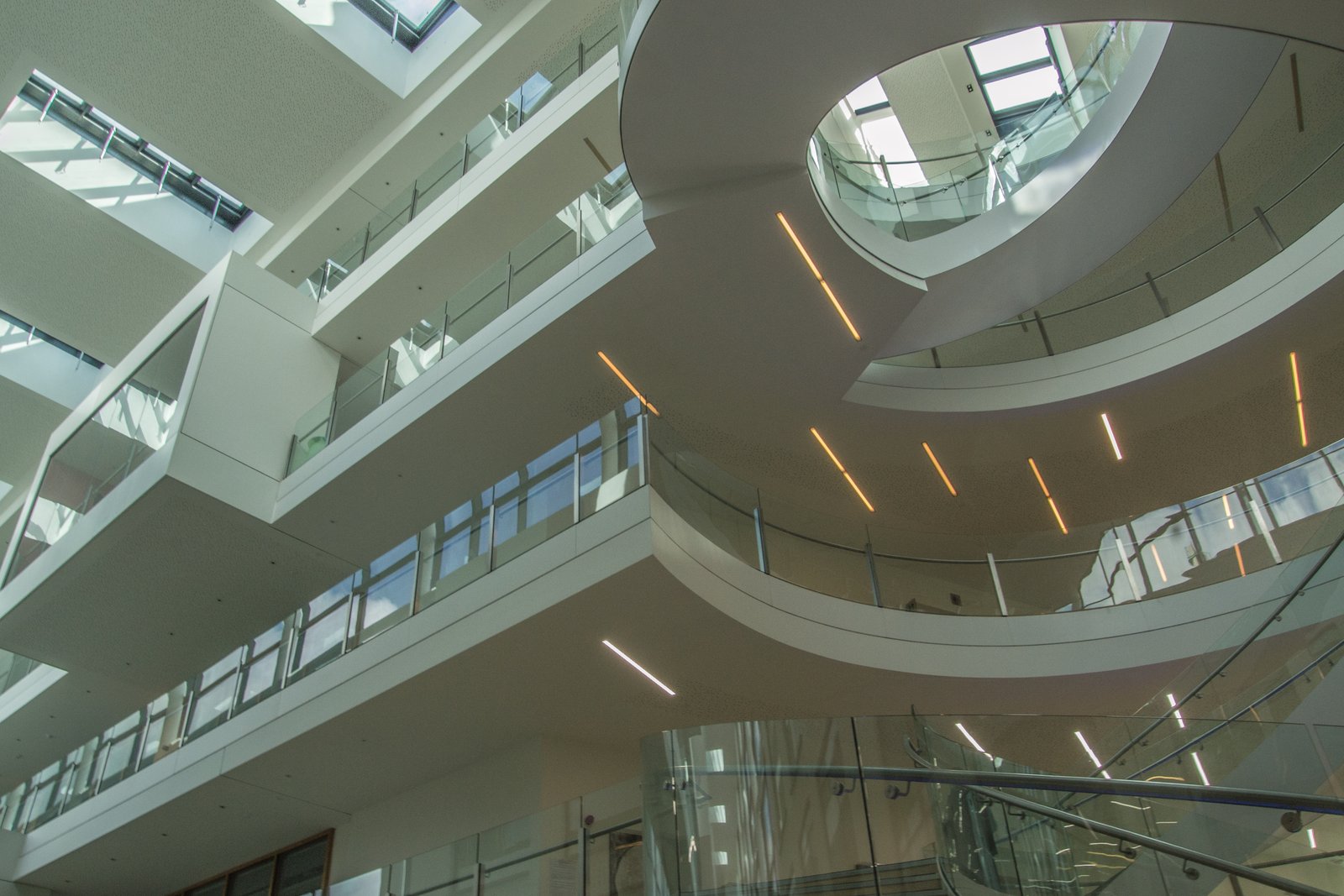 An atrium seen from below: white cantilevered galleries with glass balustrades step up around a curved drum of balconies, warm strip lights set into the soffits, a glass-sided spiral stair rising at the lower right, and daylight coming through skylights above.