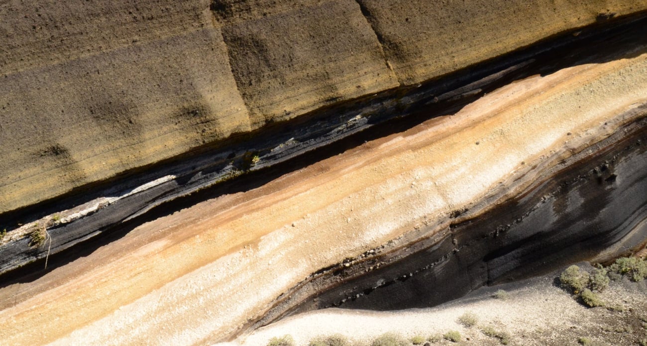 Tilted volcanic strata filling the whole frame: bands of dark tephra, rust, ochre and pale pumice running diagonally, each layer a separate eruption laid down and buried by the next.